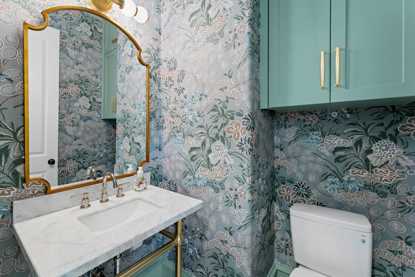 Powder room with floral wallpaper, brass mirror, and marble console sink at Revere Pl. in Lakewood